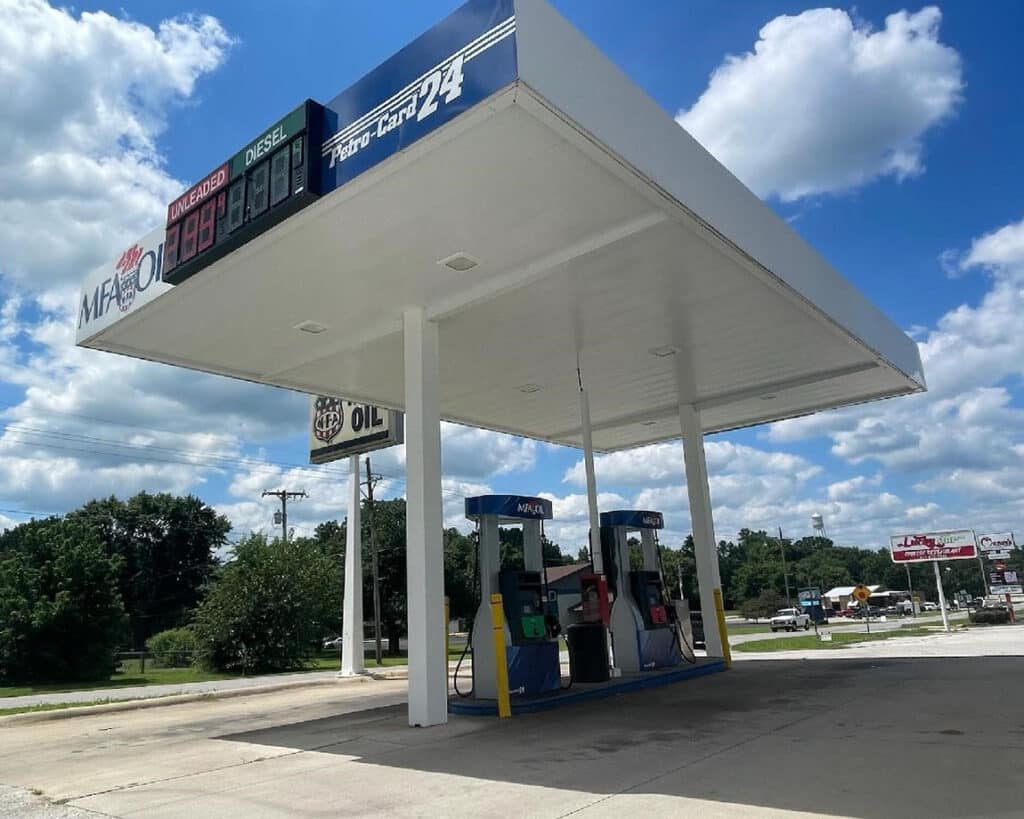 A gas station with two fuel pumps under a large white canopy displaying fuel prices. The background shows a few trees, clouds in a blue sky, and some roadside signs. The station appears quiet and empty.