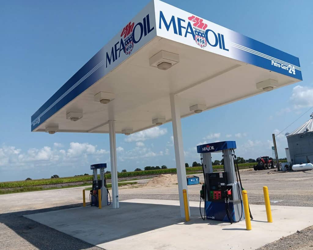 A gas station featuring two fuel pumps under a large canopy with the MFA Oil logo. The background includes a clear blue sky and fields, suggesting a countryside setting. Yellow bollards are positioned around the pumps.