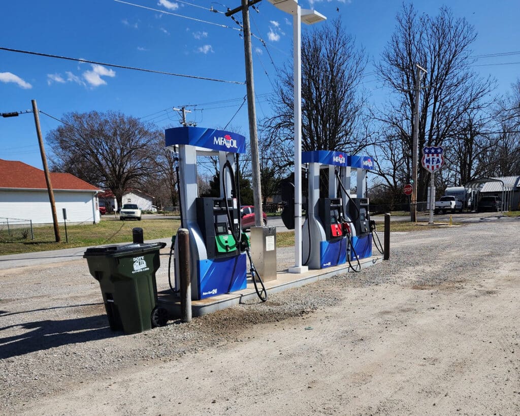 Two gasoline pumps with green and red nozzles are set in a rural area. A trash bin is nearby, and a sign reads Metro above the pumps. Trees and a small building are in the background under a clear sky.