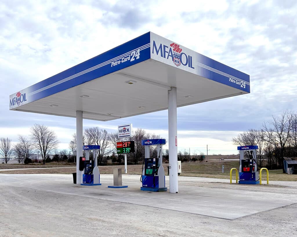 A gas station with two pumps under a canopy displaying MFA Oil and Petro-Card 24 logos. The price sign shows unleaded at $2.87 and premium at $3.39. The area is surrounded by trees and open sky.