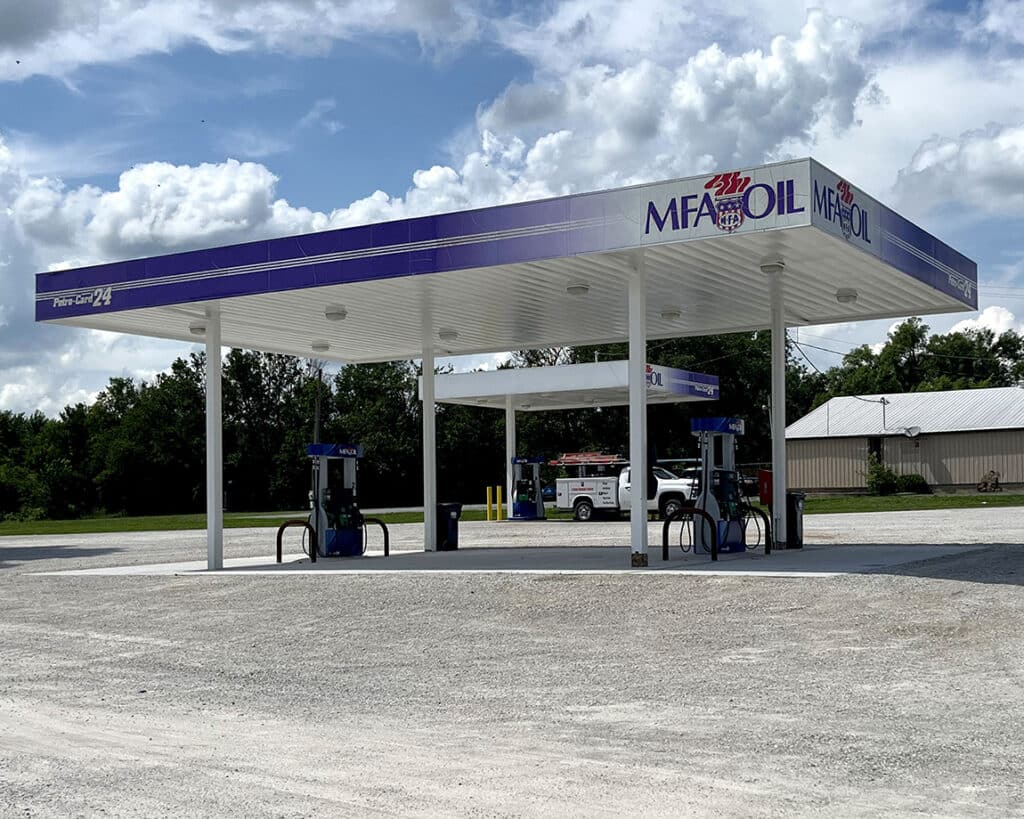 A small, empty MFA Oil gas station with two pumps sits on a gravel lot under a partly cloudy sky, surrounded by trees and a building in the background.