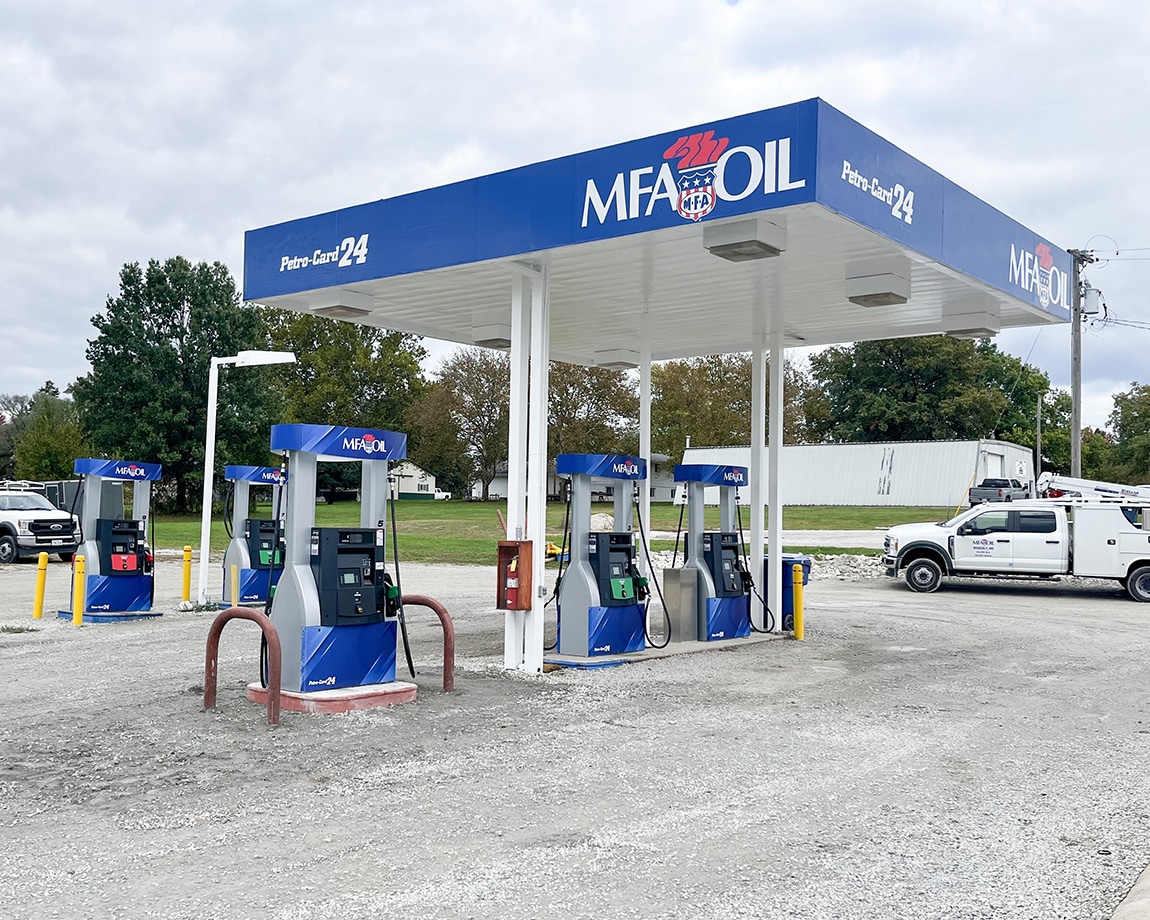 A small MFA Oil gas station with four fuel pumps under a blue and white canopy on a gravel lot; a white pickup truck is parked nearby, and trees and a white building are in the background.