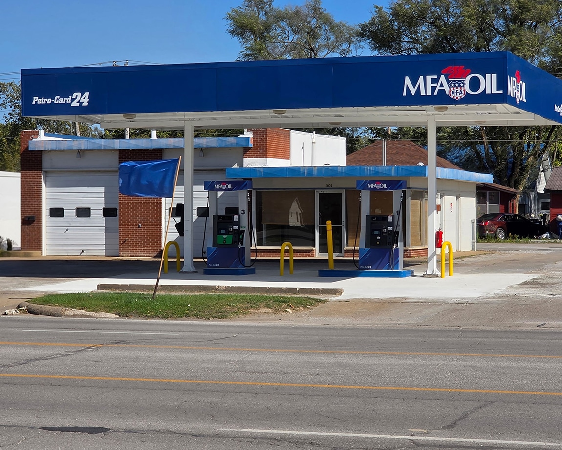 A small MFA Oil gas station with two fuel pumps under a blue canopy stands near a road. The station has a white and brick building, garage doors, and minimal landscaping. Trees and houses are visible in the background.