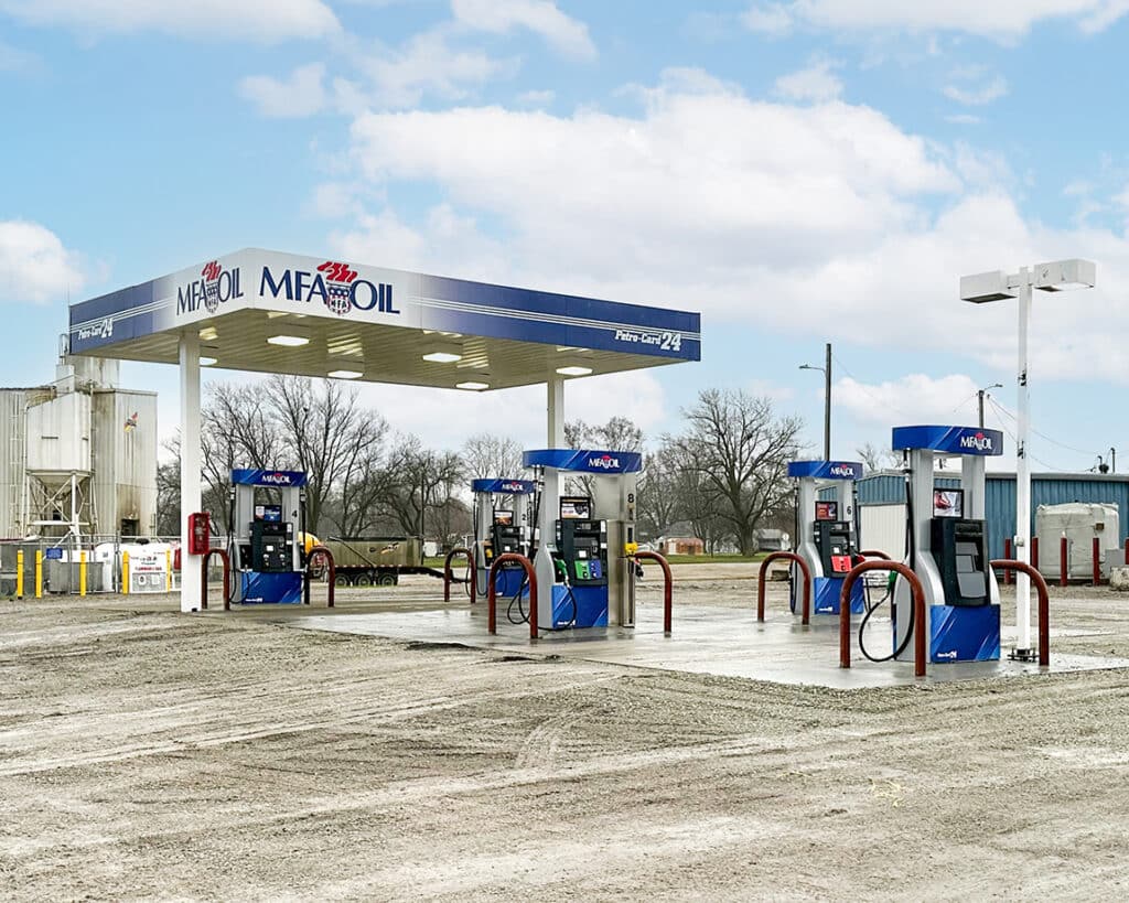 A small MFA Oil gas station with four fuel pumps under a blue and white canopy, set on a concrete island, surrounded by bare trees and a dirt lot under a cloudy sky.