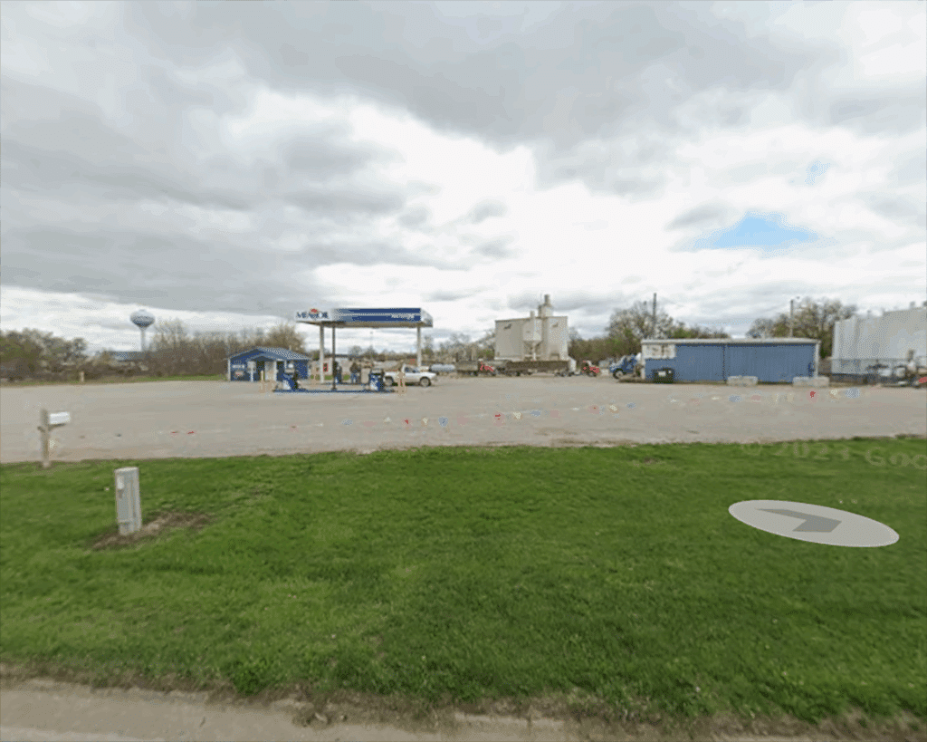 A small gas station with a blue-and-white canopy sits in a large, mostly empty parking lot. There are a few vehicles parked. Grass and a road are in the foreground under a cloudy sky.