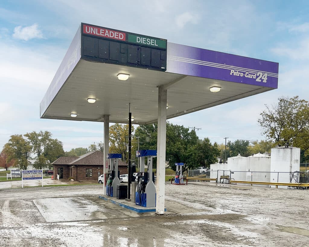 A small, rural gas station with a purple and white canopy labeled Petro-Land 24. The fuel pumps are empty and the price signs for unleaded and diesel are blank. The ground is wet, and there are trees and a building in the background.