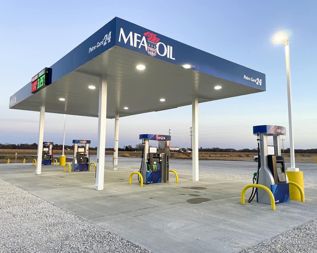 A clean, well-lit MFA Oil gas station with four fuel pumps stands on a concrete lot beside a gravel area, surrounded by open fields under a clear sky at dusk.