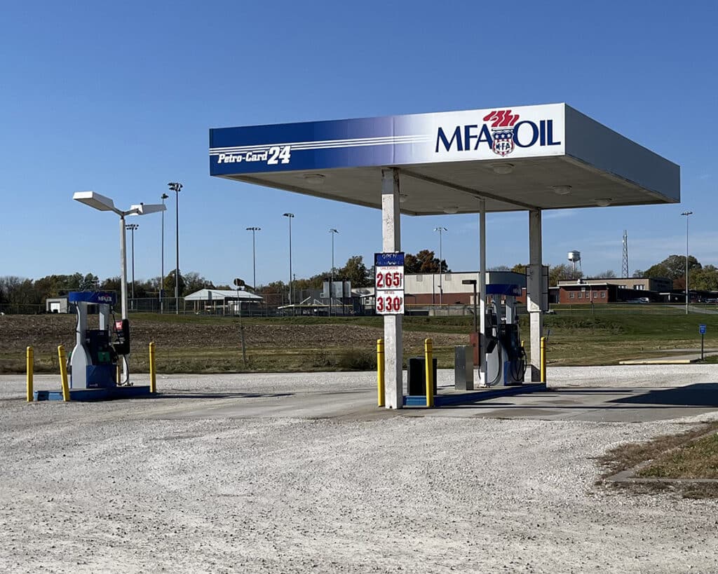 A small MFA Oil gas station with two fuel pumps under a canopy, displaying fuel prices of $2.65 and $3.30. The station is in a rural area with open land and buildings in the background.