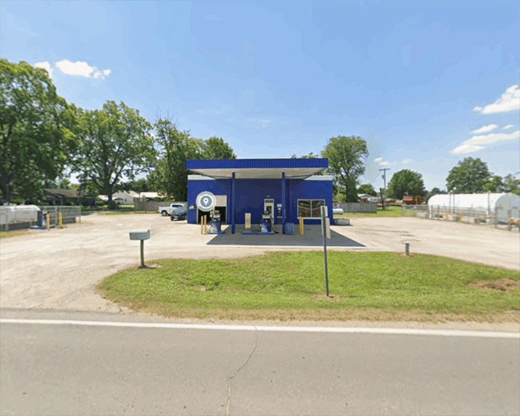 A small blue gas station stands alone on a sunny day, surrounded by a large empty lot with trees and some buildings in the background. There is a road in front of the station and a single vehicle parked beside it.