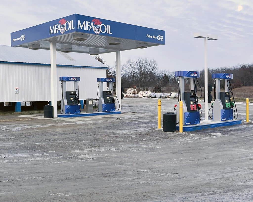 A rural gas station with three blue MFA Oil fuel pumps under a blue canopy next to a white building, surrounded by an empty, slightly muddy lot and leafless trees in the background.