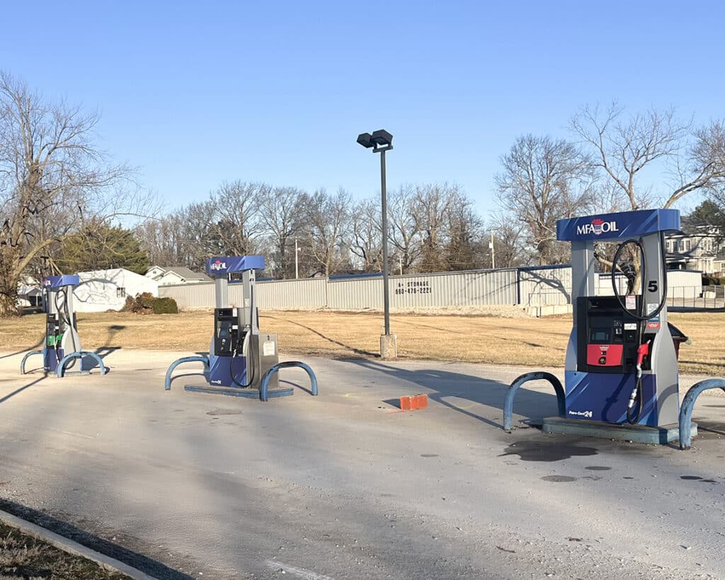 Three blue and white gas pumps stand on a mostly empty concrete lot under a clear sky. The area is surrounded by dry grass, leafless trees, and a metal fence in the background.