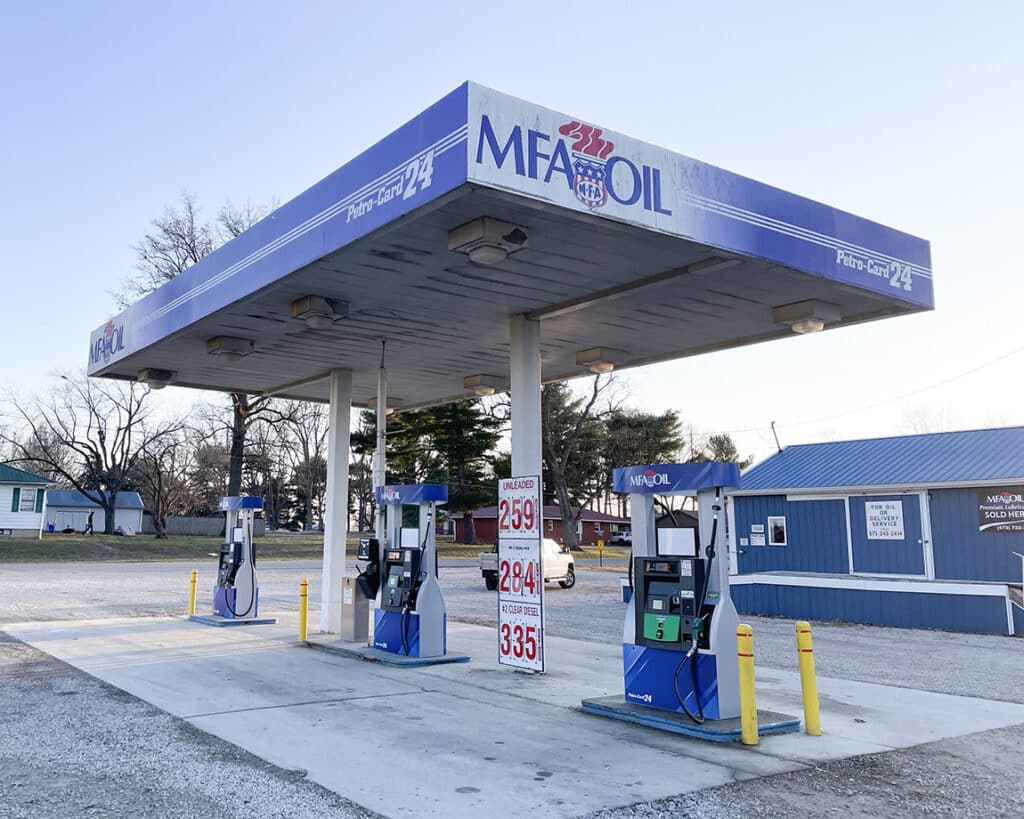 A small MFA Oil gas station with two pump islands, a blue canopy, and posted fuel prices. The background includes a blue building, trees, and a clear sky.