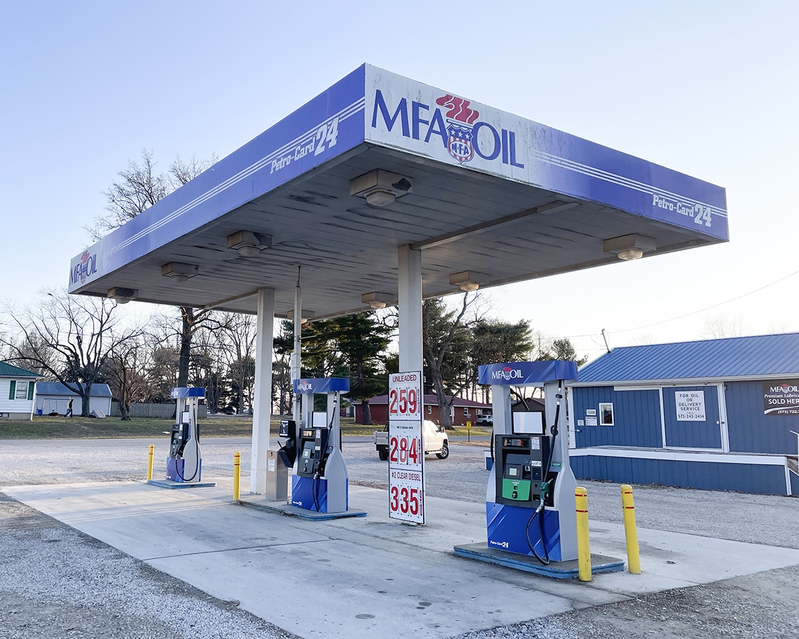 A small MFA Oil gas station with two pump islands, a blue canopy, and posted fuel prices. The background includes a blue building, trees, and a clear sky.