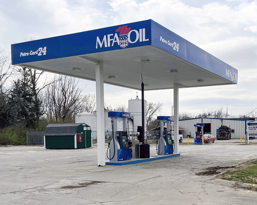 A small outdoor MFA Oil gas station with two blue and white fuel pumps under a white canopy. A green dumpster, trees, and a metal building are visible in the background on a cloudy day.