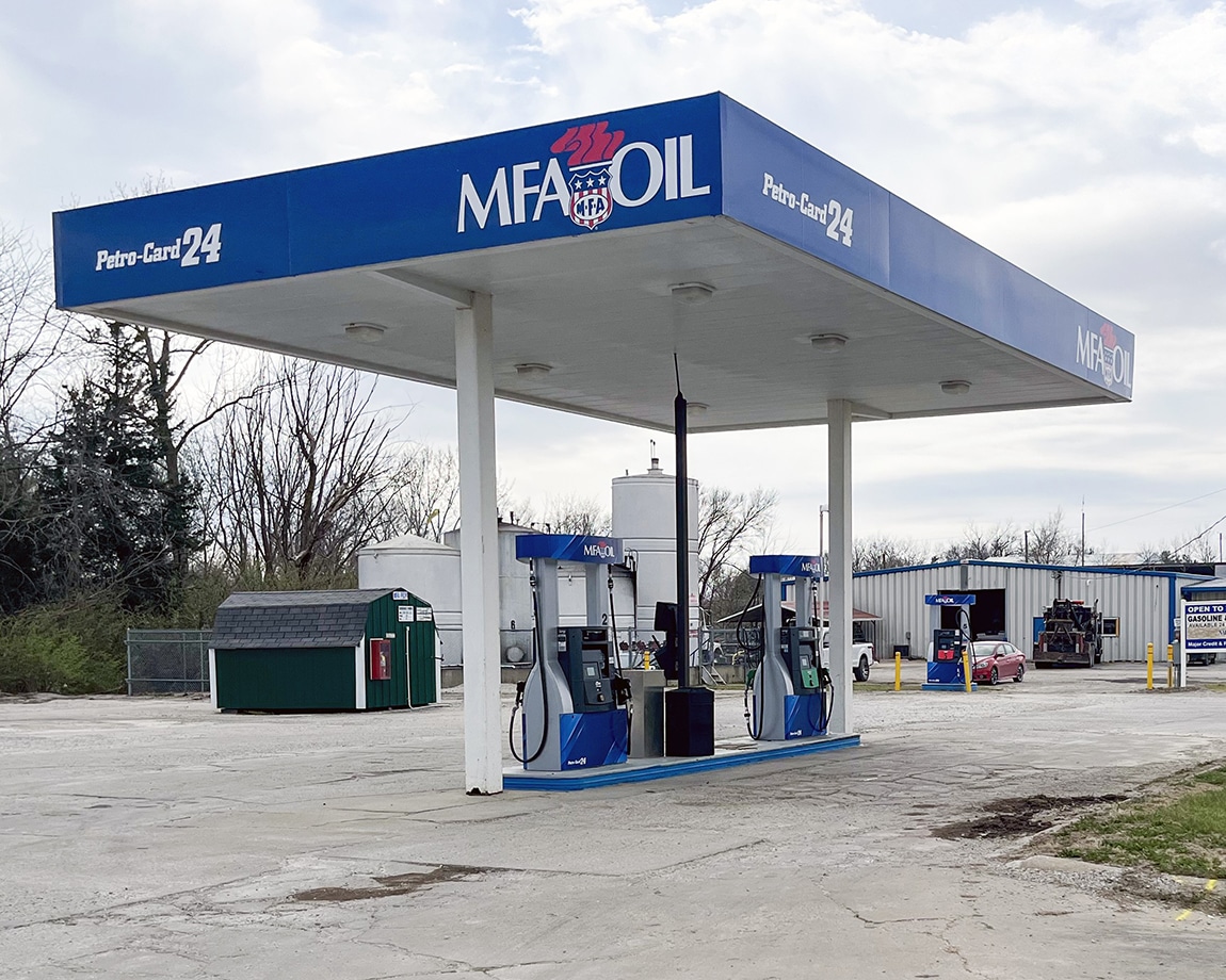 A small outdoor MFA Oil gas station with two blue and white fuel pumps under a white canopy. A green dumpster, trees, and a metal building are visible in the background on a cloudy day.